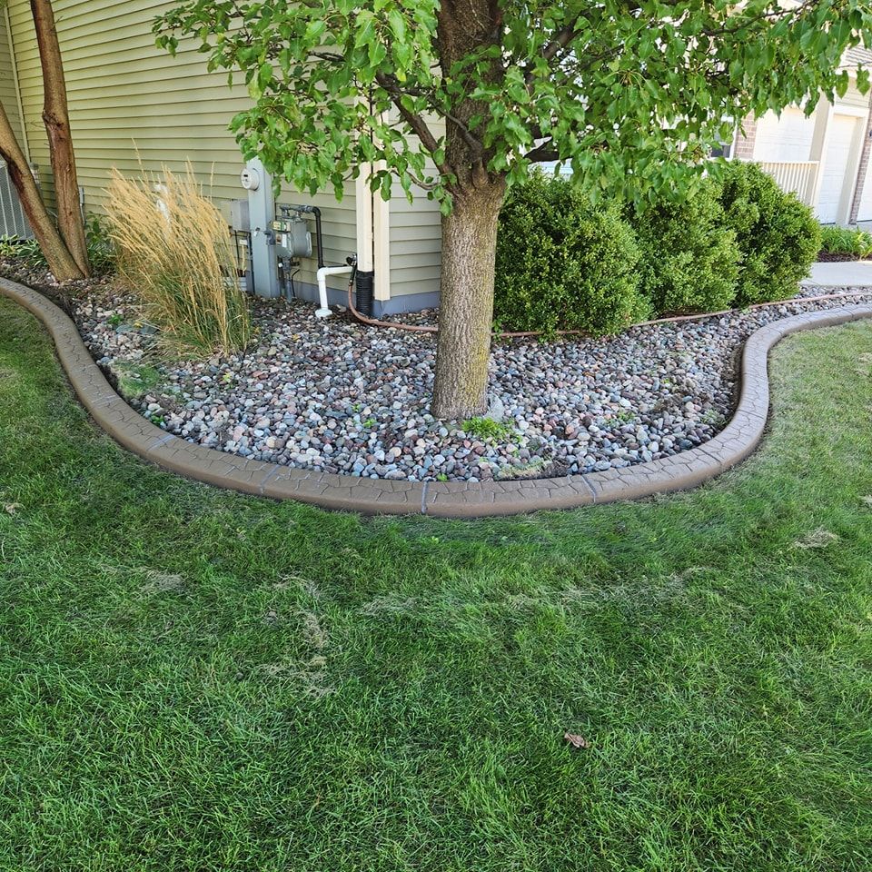 A brown, curved landscape border separates a grassy lawn from a tree and stone mulch garden bed against a house wall.