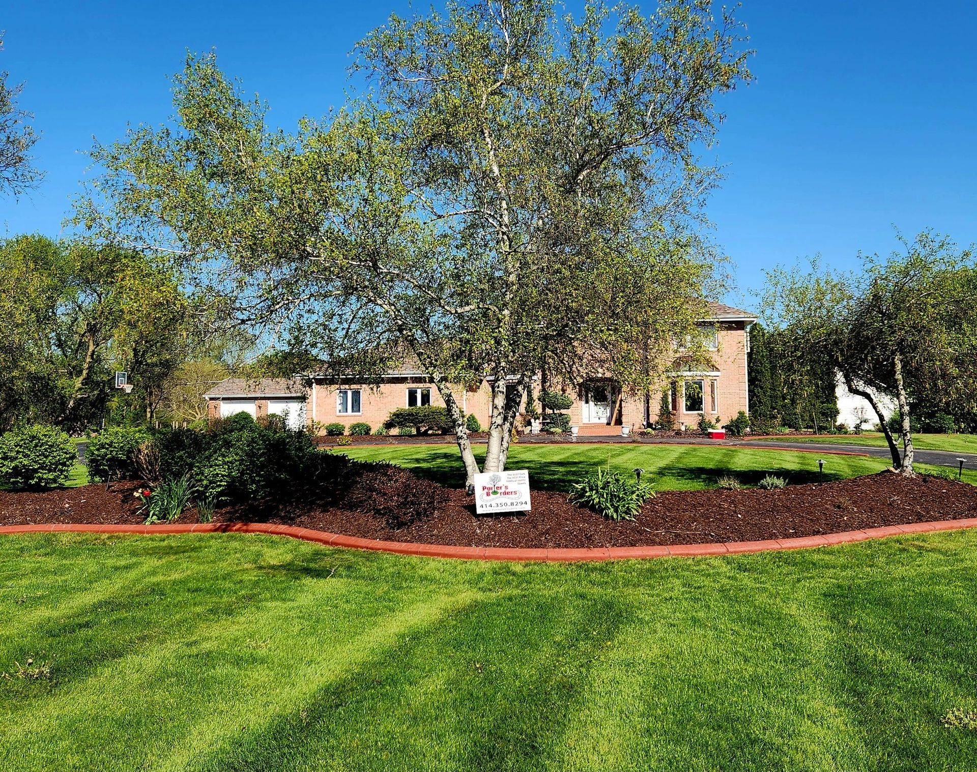 A two-story brick house sits behind a landscaped front yard with a large tree, mulch beds, and striped green grass.