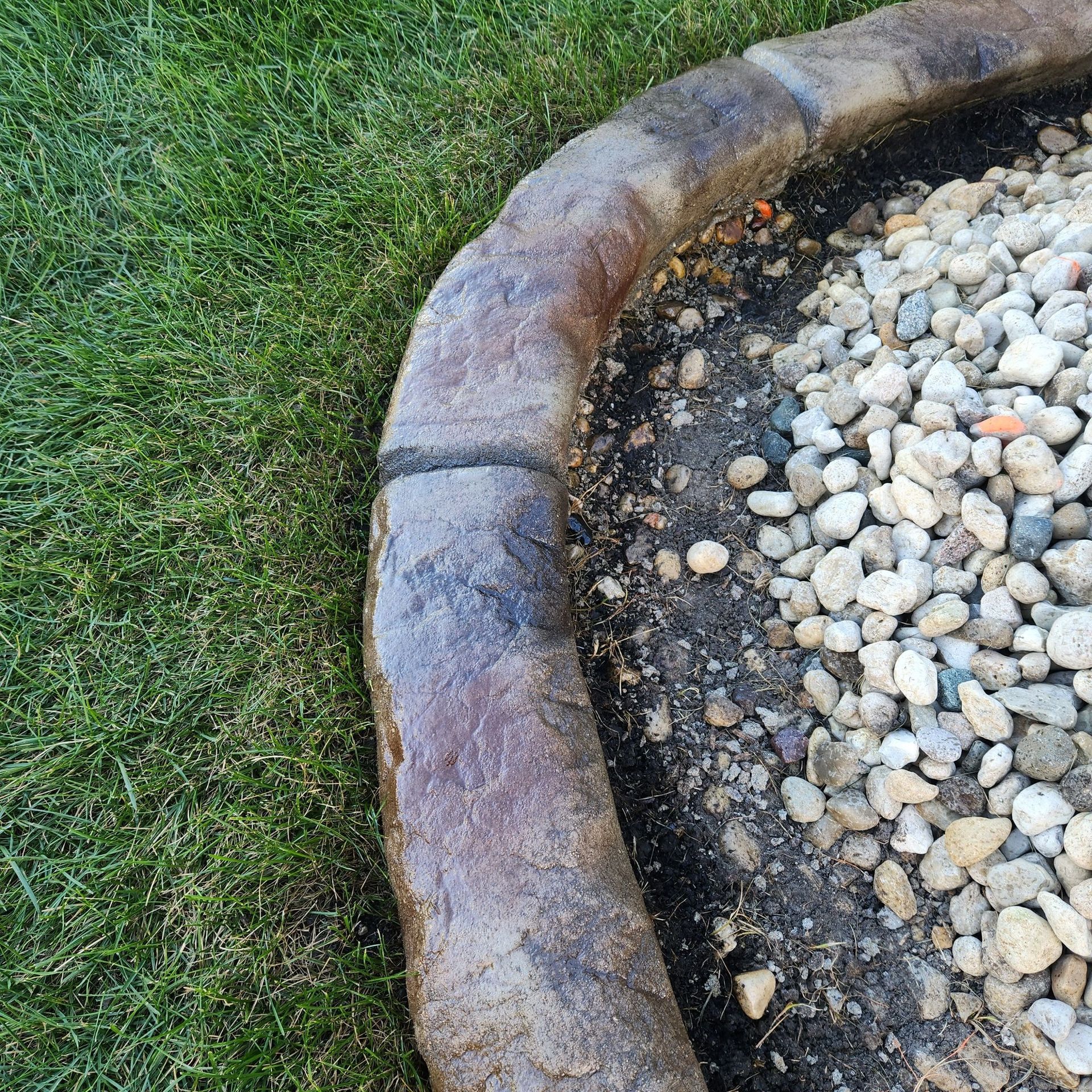 A curved landscape border of textured brown concrete blocks separating green grass from a bed of grey garden pebbles.
