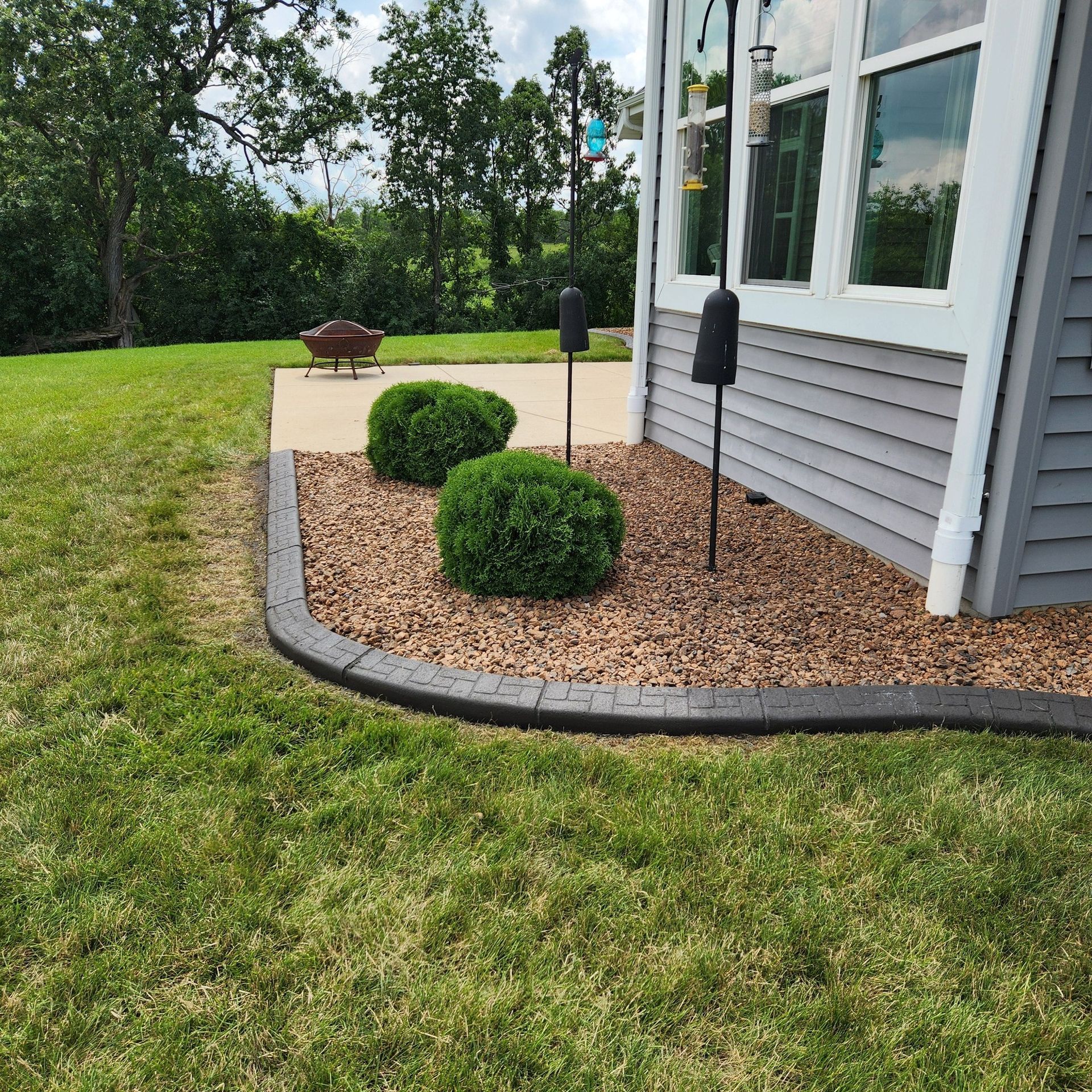 A house corner with tan siding and a landscaped garden bed containing two round shrubs, stone mulch, and gray curbing.