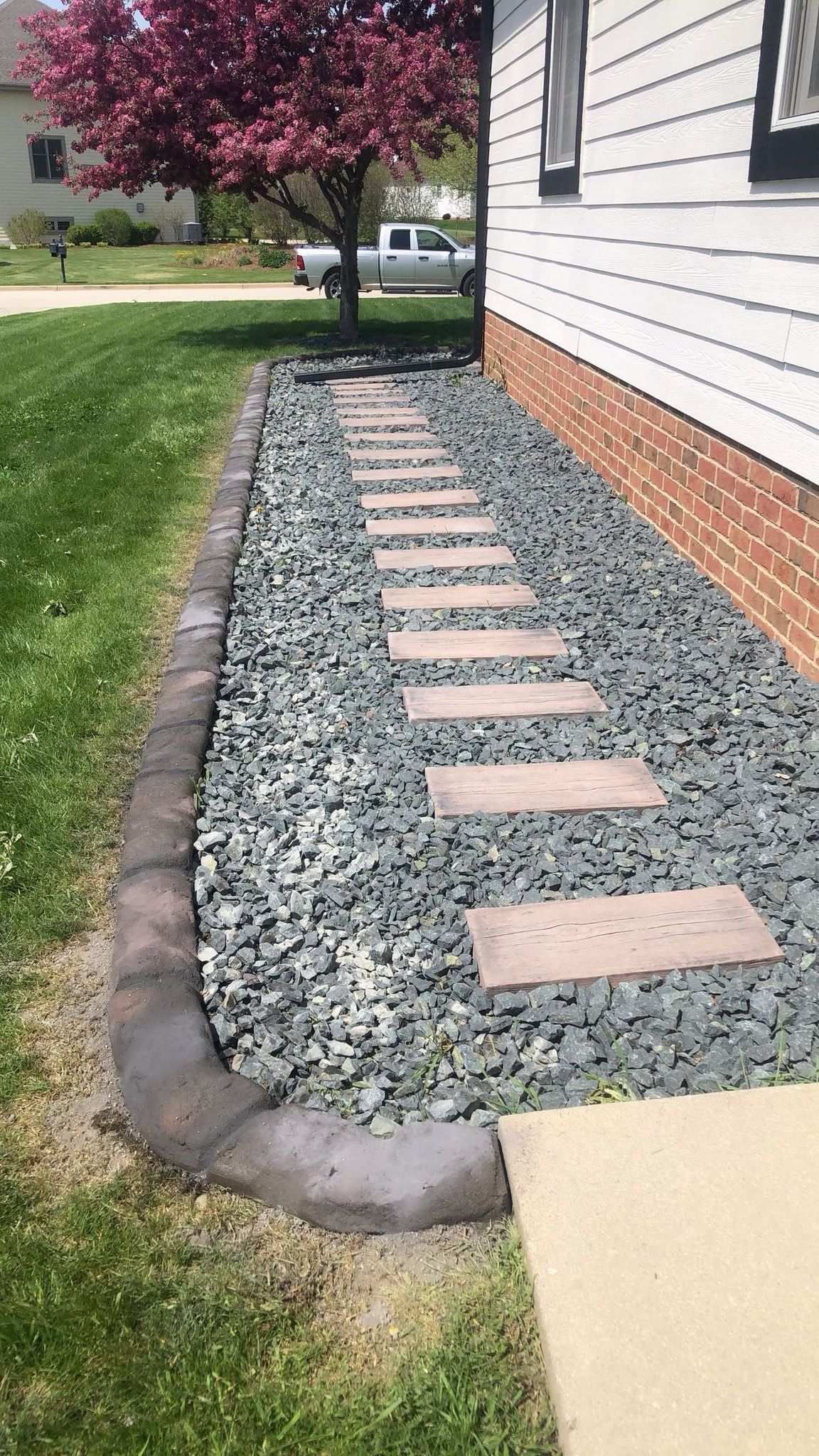 A stone path with rectangular stepping stones set in gray gravel alongside a white house with a brick foundation.