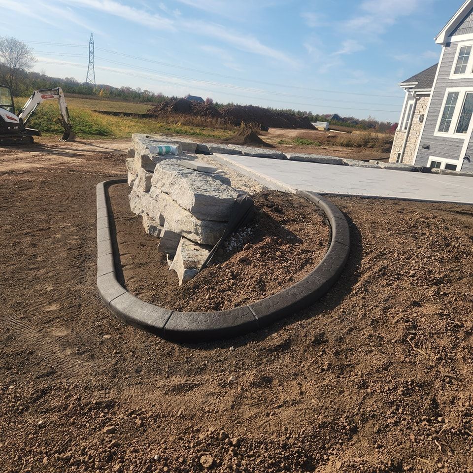 A landscaped yard under construction, featuring large grey boulders partially enclosed by a curved dark concrete edging.