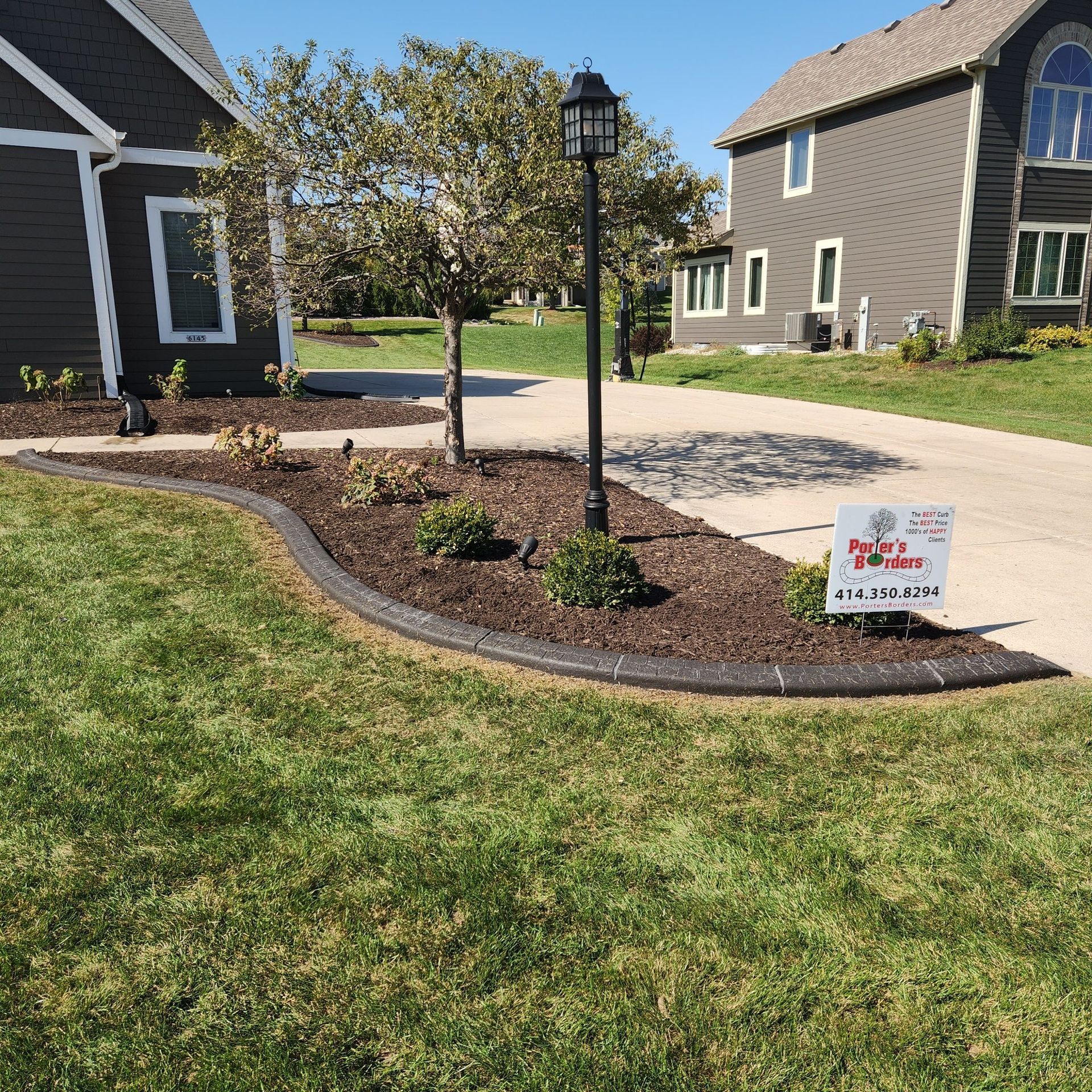 A concrete landscape border separates a mulched garden bed from a lawn in front of two suburban homes.