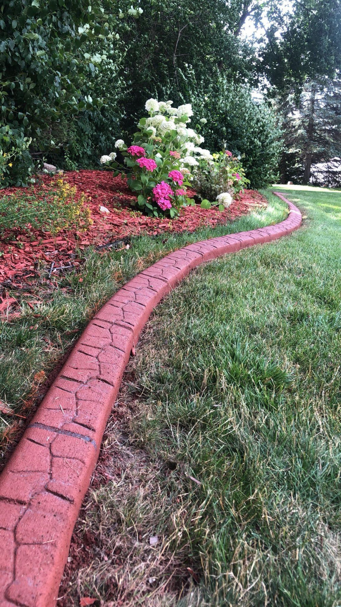 A decorative red stamped concrete landscape border separates a mulched garden bed with flowers from a grass lawn.