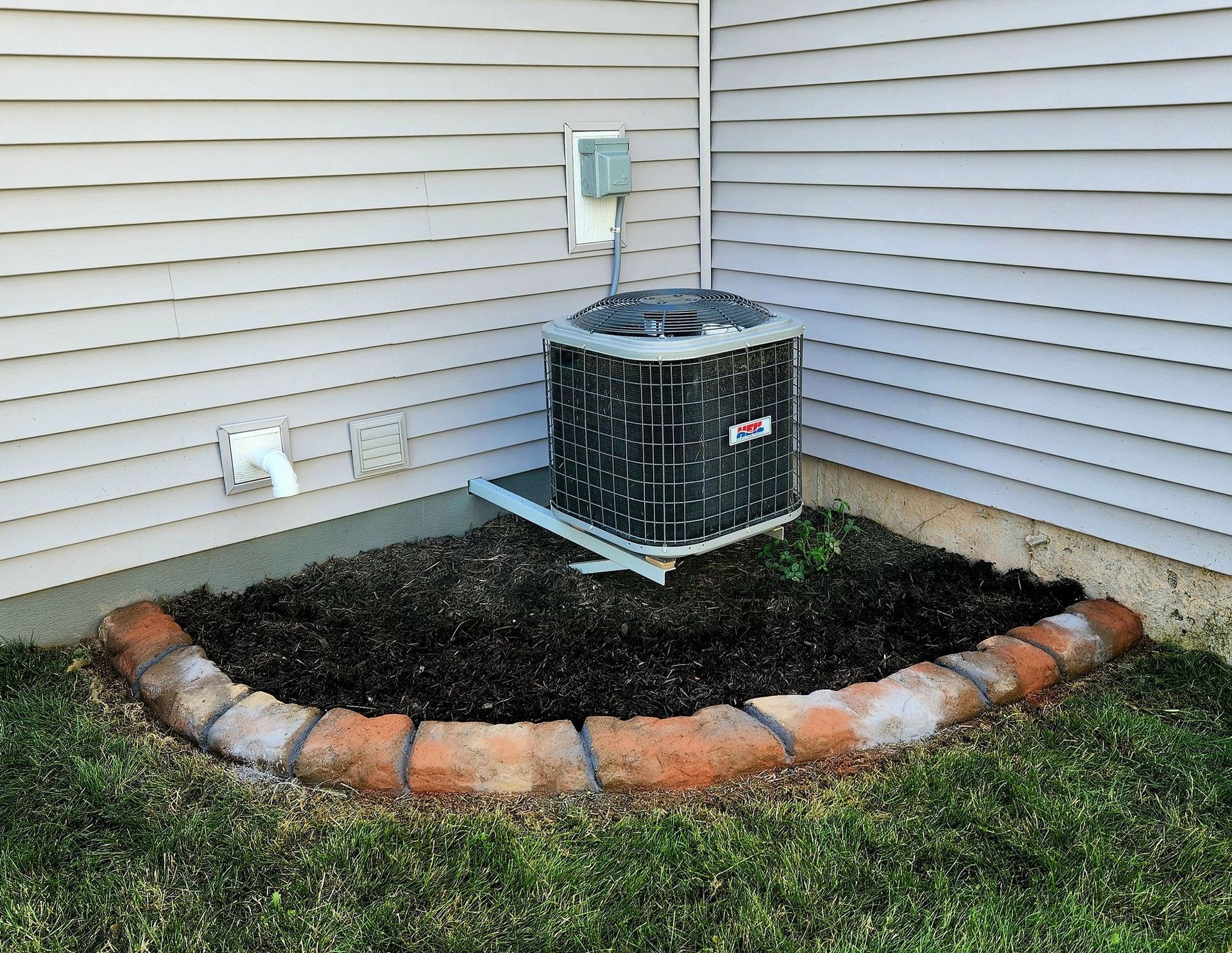 An outdoor air conditioning unit sits on a metal stand against the corner of a house, surrounded by a curved brick border.