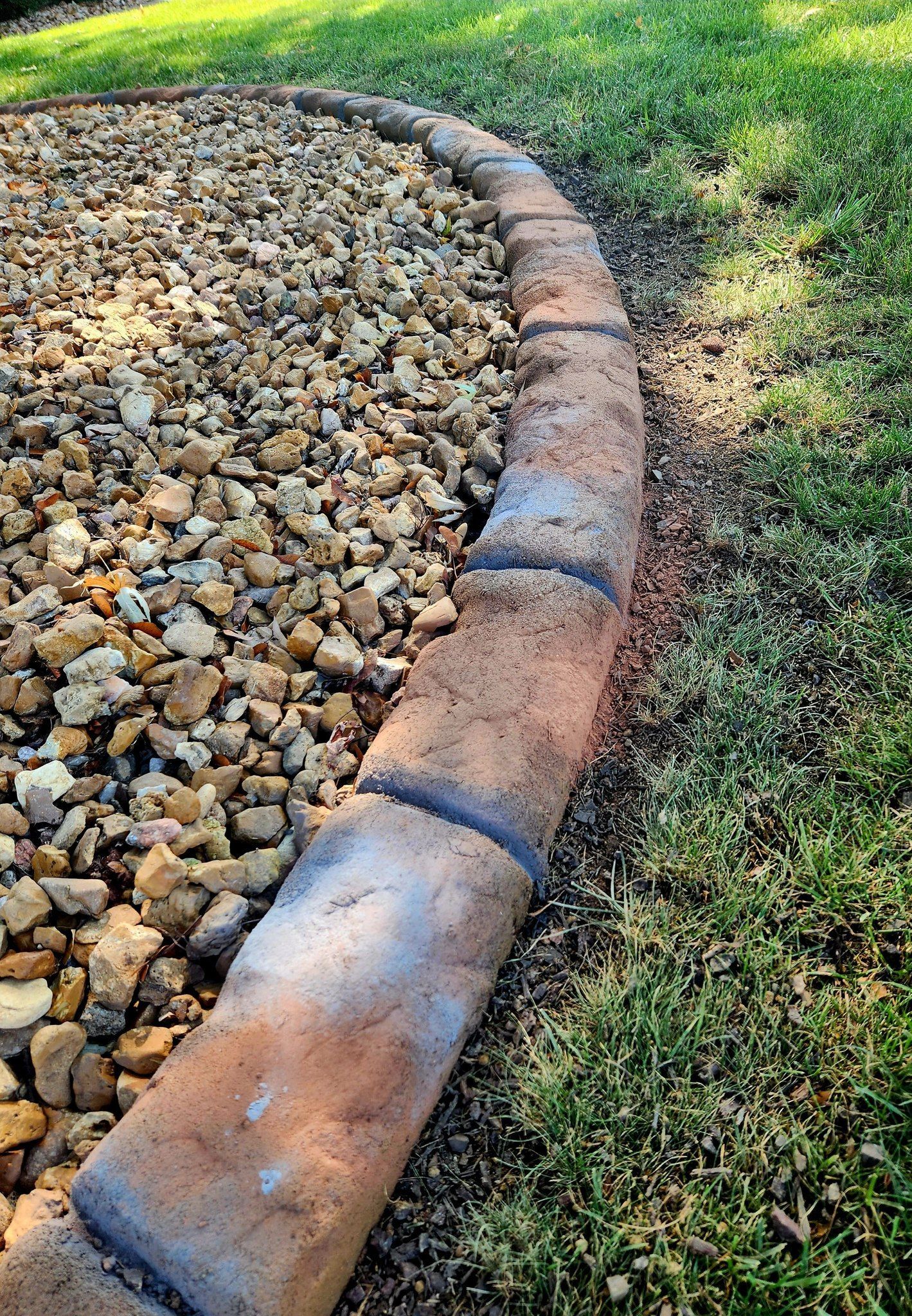 A curved stone landscape border separates a bed of small rocks from a grassy lawn.