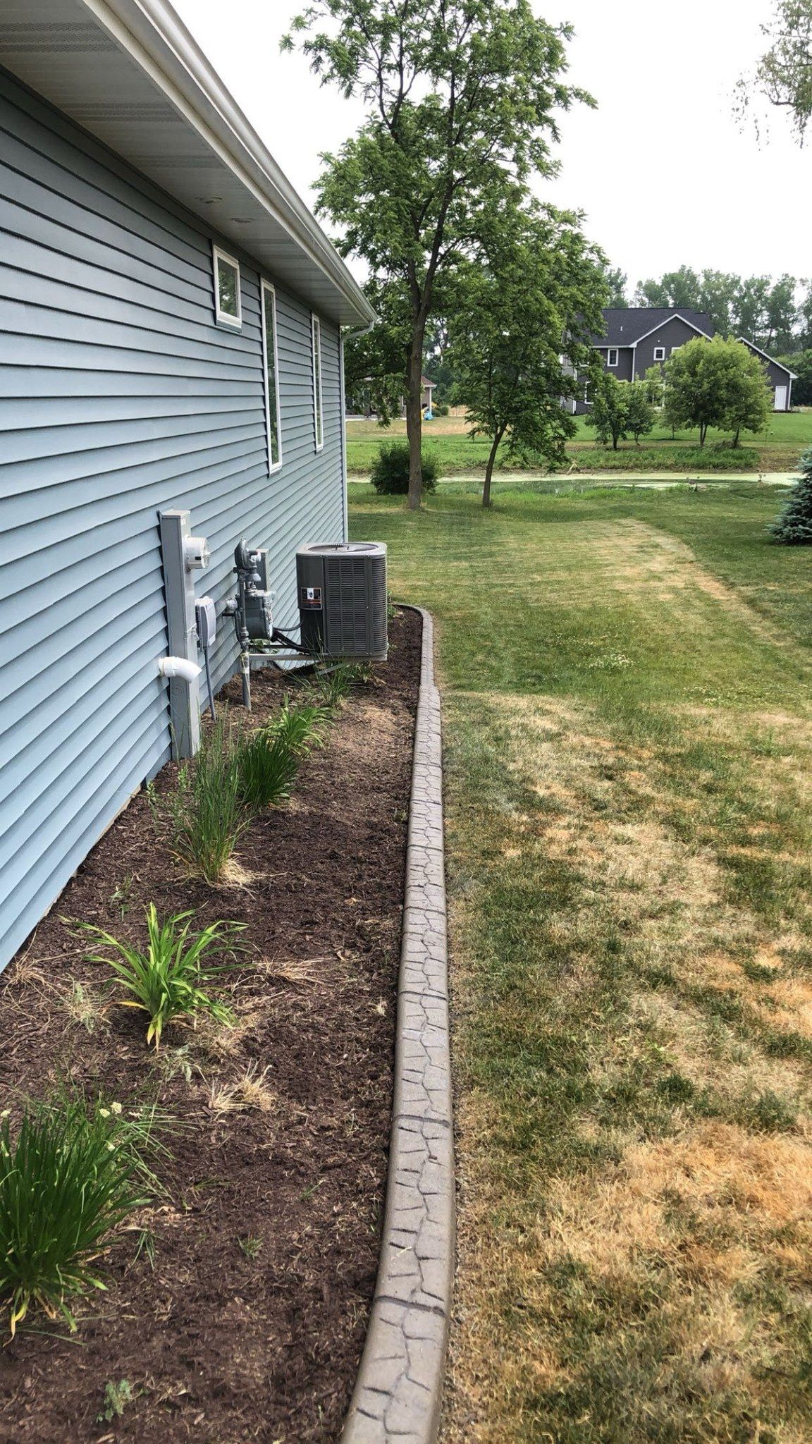 A blue-sided house with a mulched garden bed and decorative concrete edging bordering a grassy lawn.