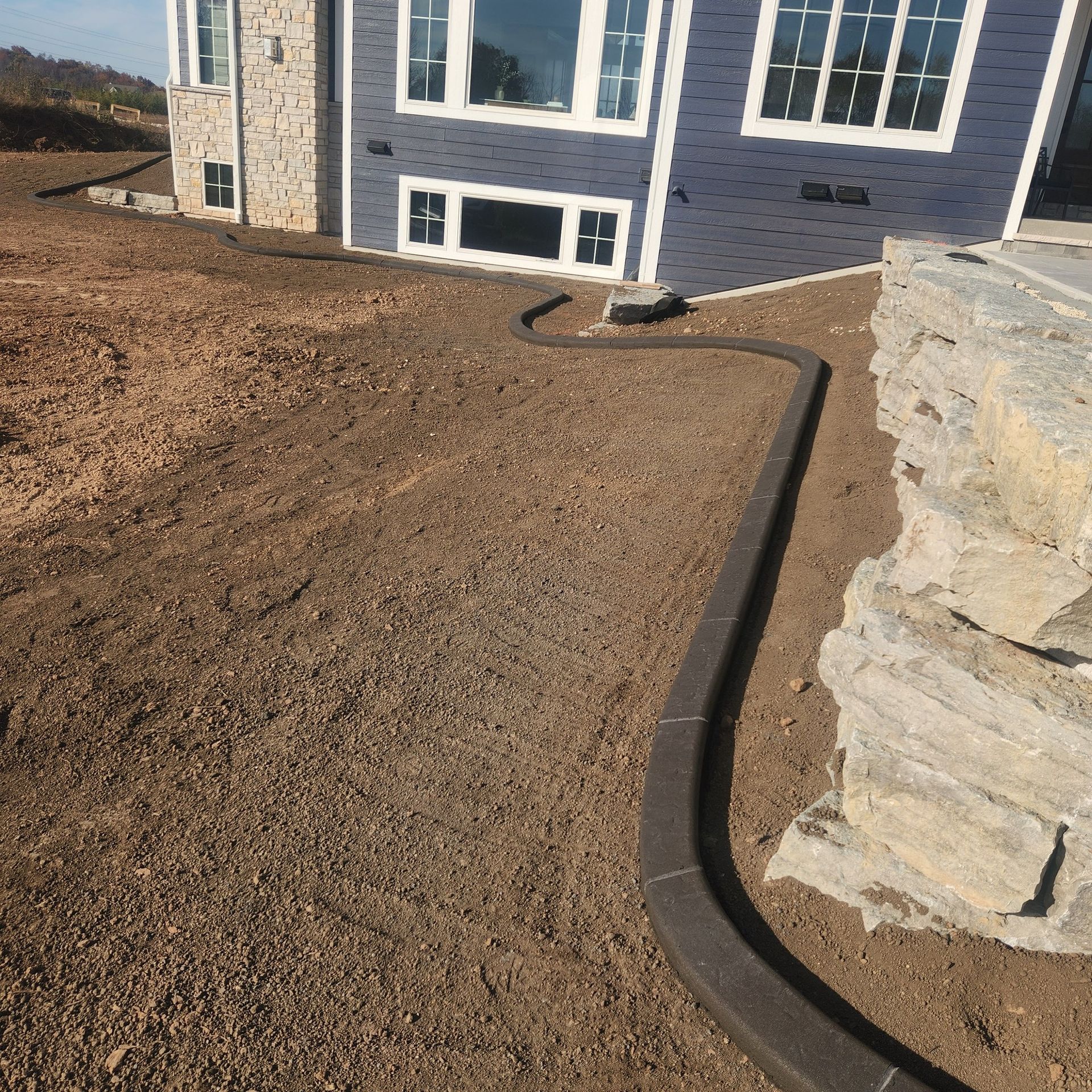 A curved concrete landscape border edges a dirt yard in front of a blue house with stone siding.