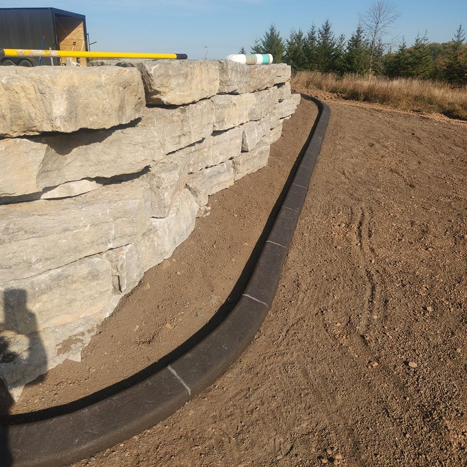 A stone retaining wall alongside a curved dark concrete border set in a dirt landscape under a clear blue sky.