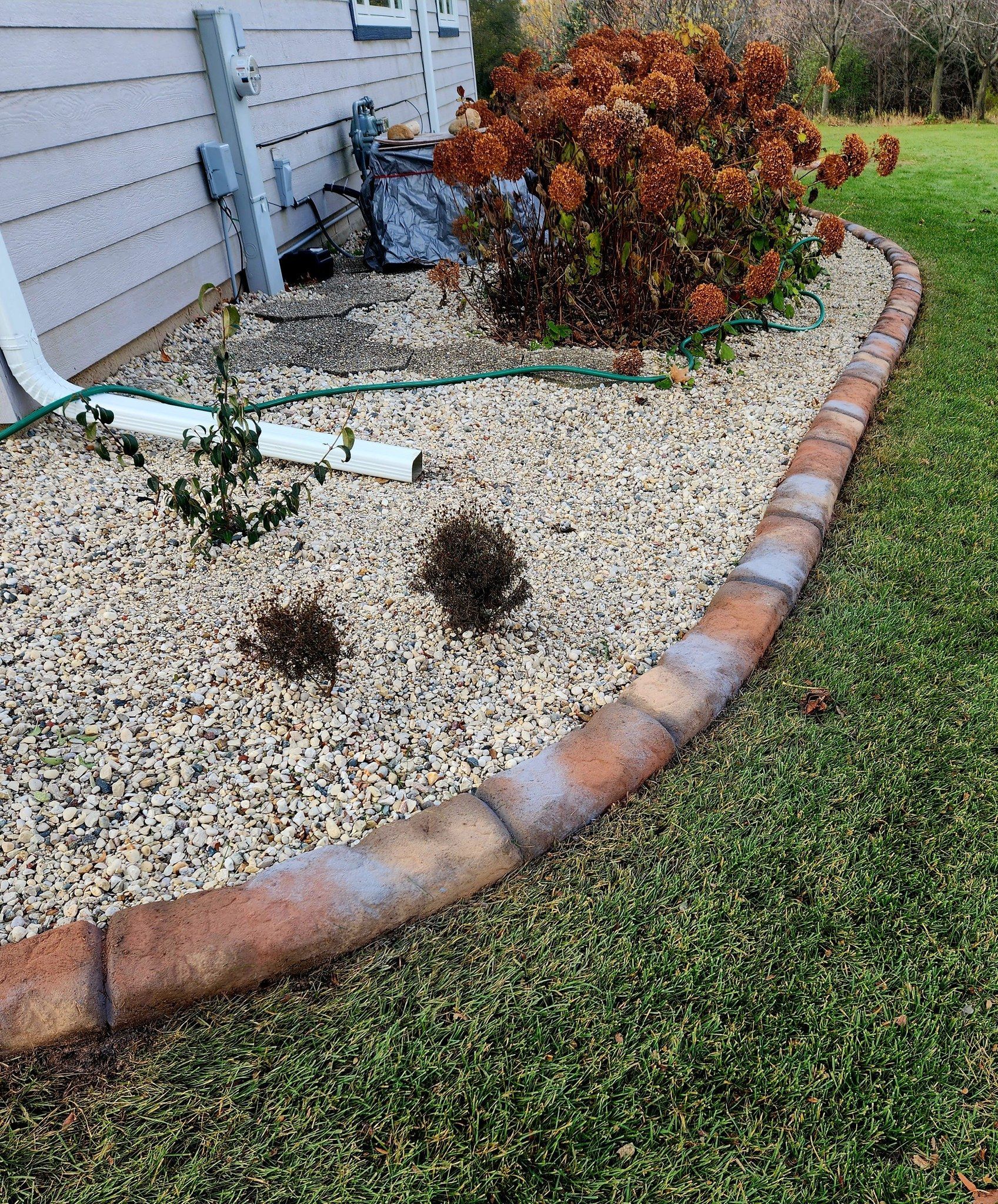 A curved row of reddish-brown concrete paver blocks borders a gravel garden bed next to a house siding and lawn.