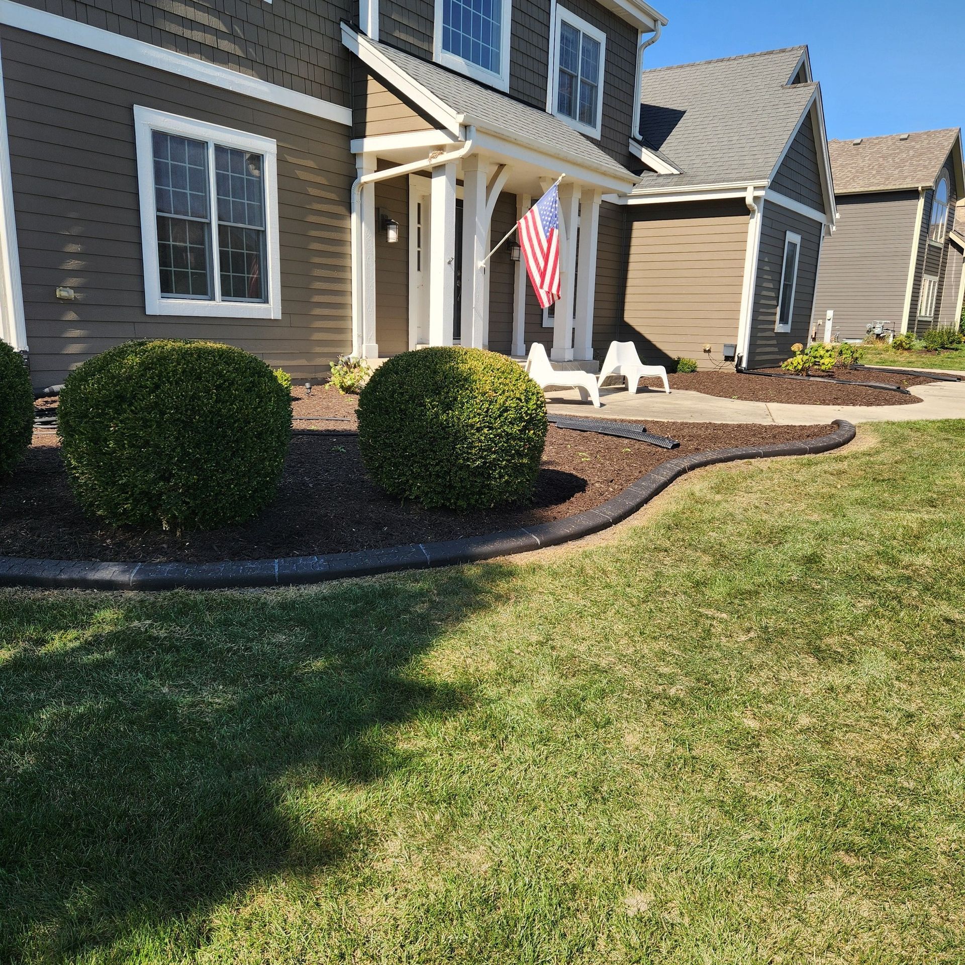 A brown suburban home with a green lawn, dark mulch flower beds, manicured shrubs, and an American flag on the porch.
