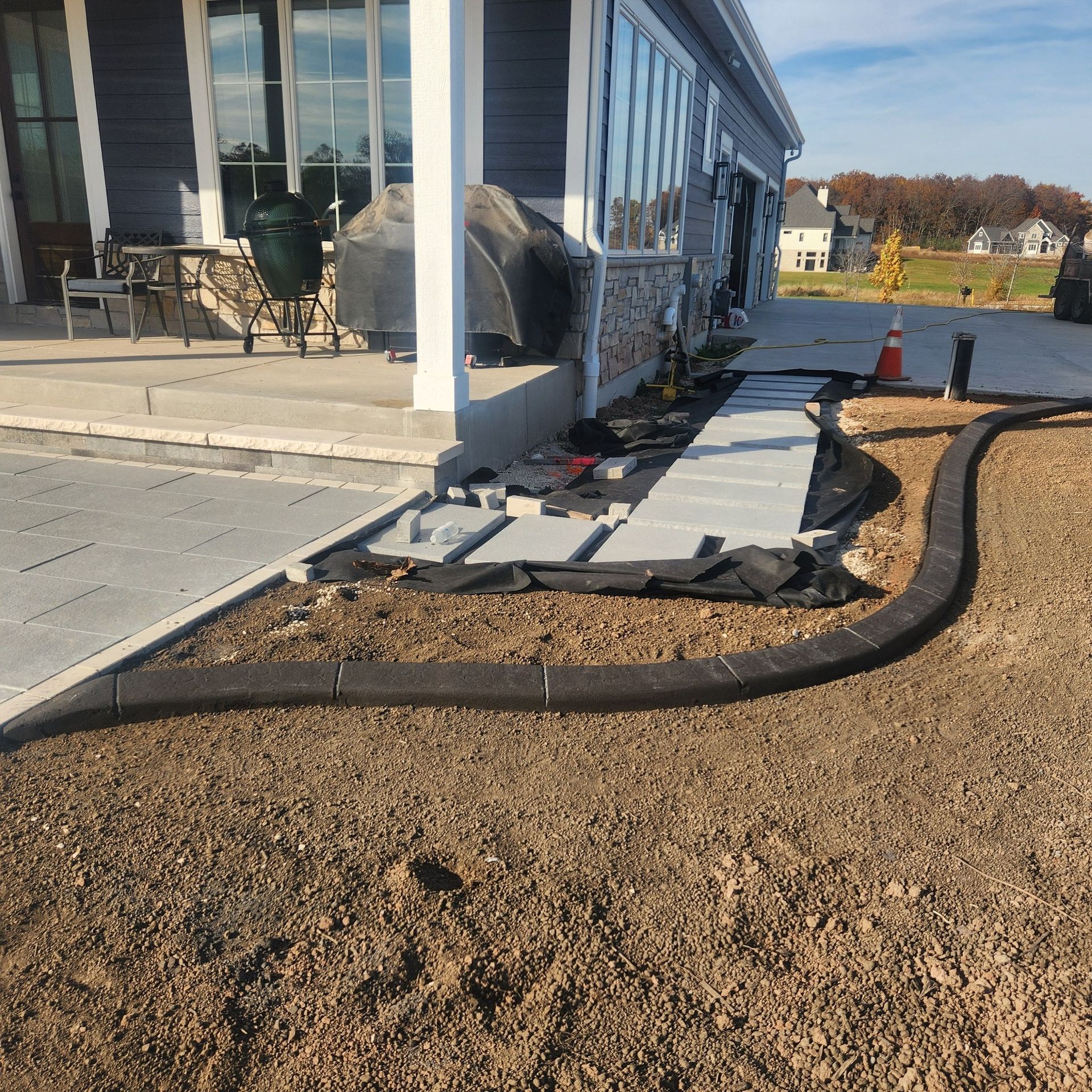 A partially finished landscape project featuring a curved concrete border, grey stone pavers, and a dirt yard by a house.