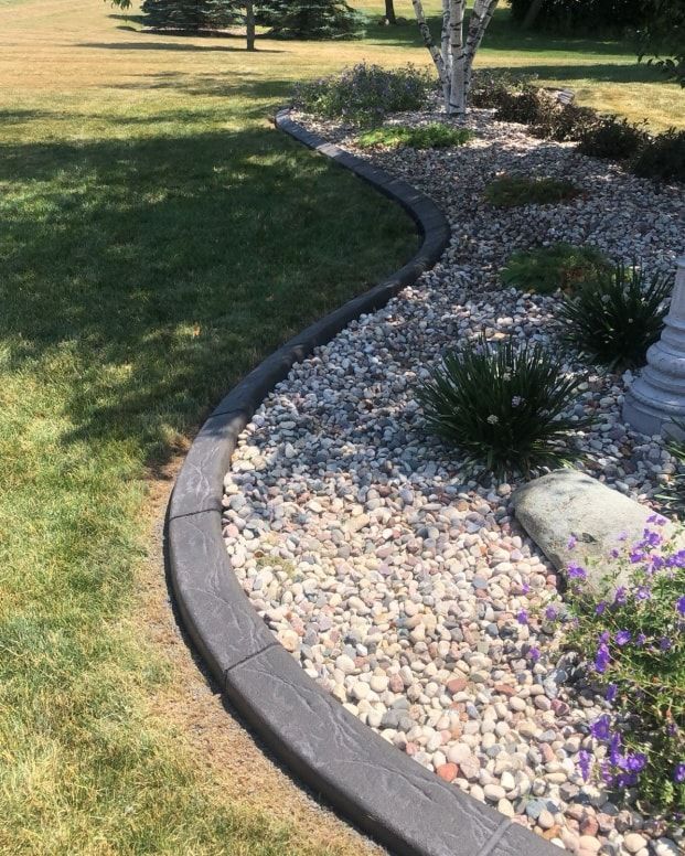 A curved landscape border made of dark wood-textured blocks separates a lush green lawn from a garden bed of light rocks.