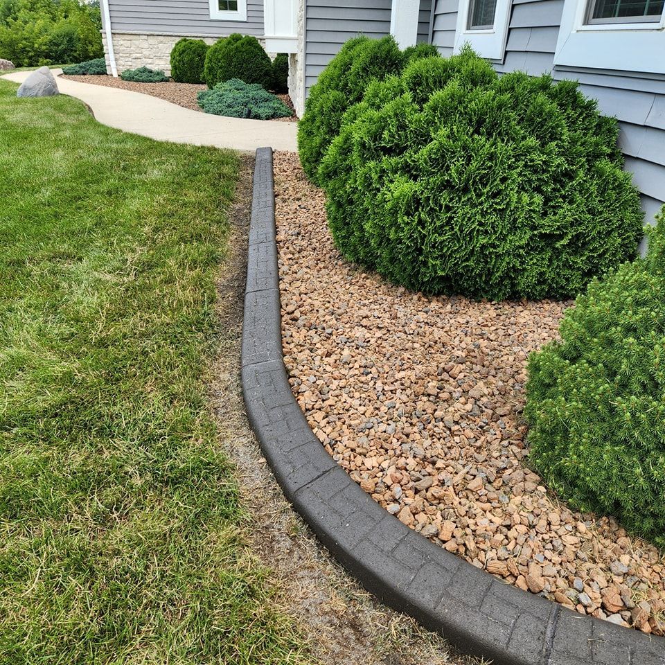 A gray stone landscape border separates a patch of green grass from a garden bed filled with brown mulch and shrubs.