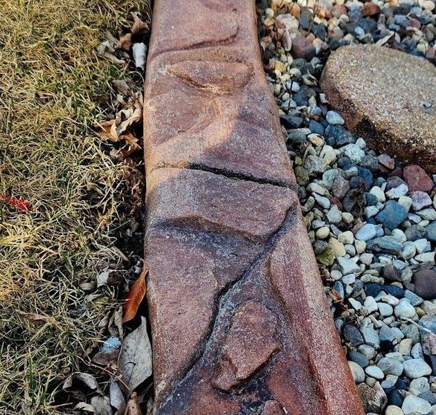 A top-down view of a textured, reddish-brown concrete landscape border separating dried grass from a gravel garden bed.