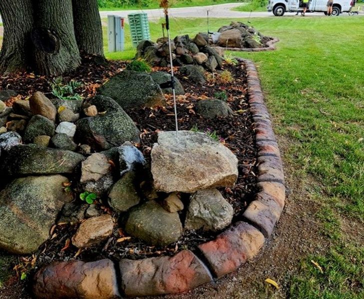 A landscaped garden bed edged with rounded, rustic-style stone pavers next to a green lawn under a large tree.