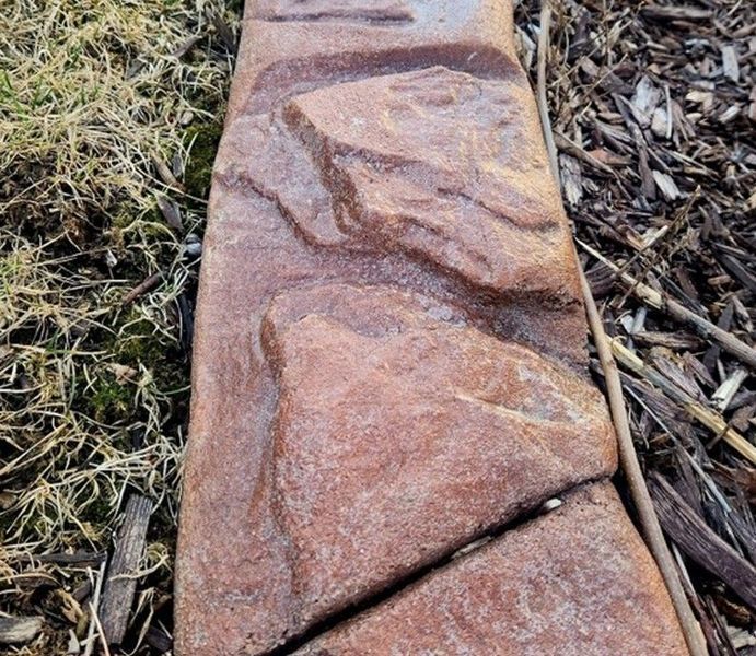 A close-up of a decorative, rust-colored concrete landscape border with a textured rock pattern separating grass and mulch.