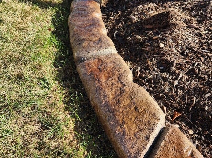 A line of textured, brown stone garden edging separates a patch of grass from a bed of wood mulch.