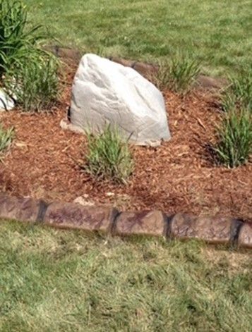 A large, light-colored decorative rock sits in a mulch bed, bordered by curved stone edging next to a green lawn.