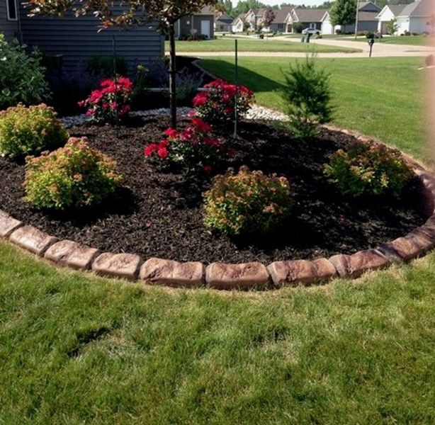 A landscaped garden bed with red roses and green shrubs, edged with stones, set in a residential lawn.