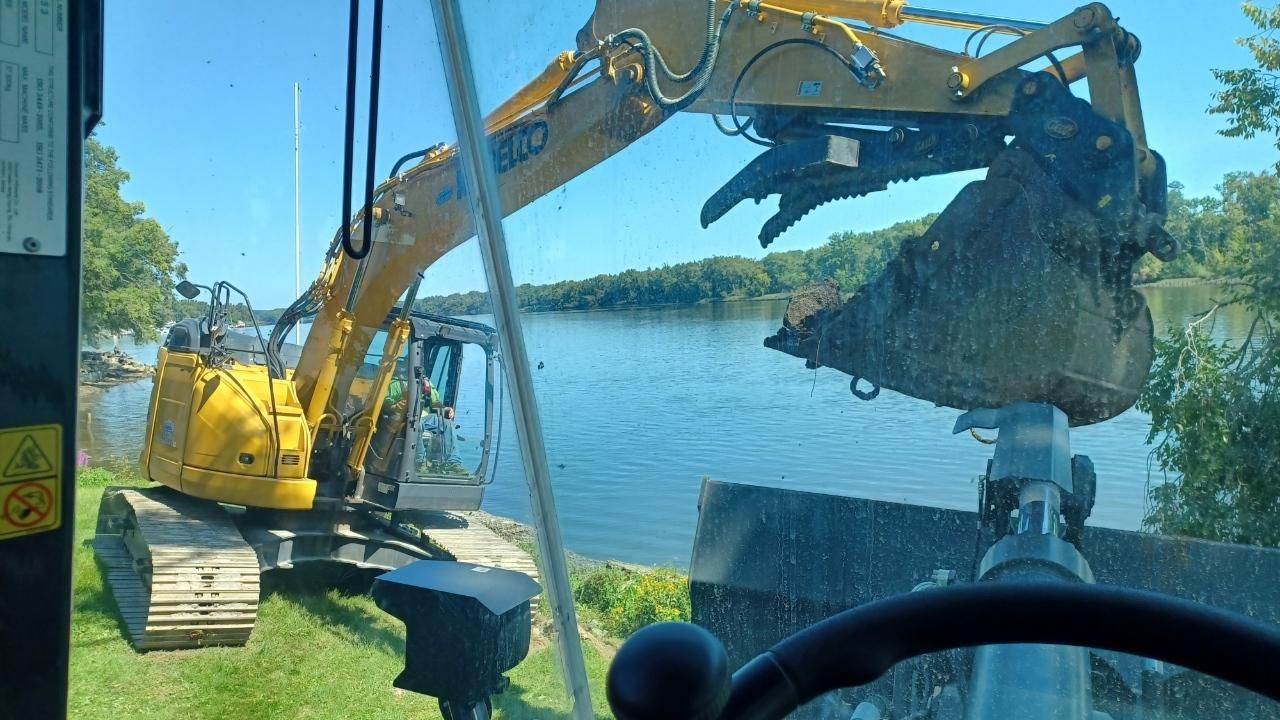 A yellow excavator is digging in the grass near a body of water.