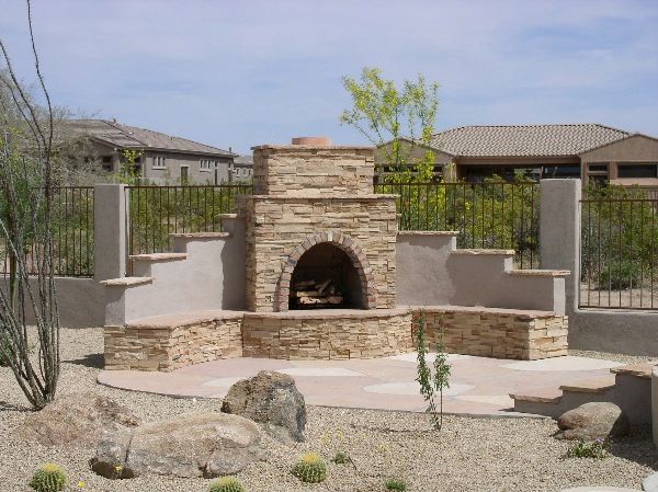A stone fireplace is surrounded by rocks and cactus in a backyard.