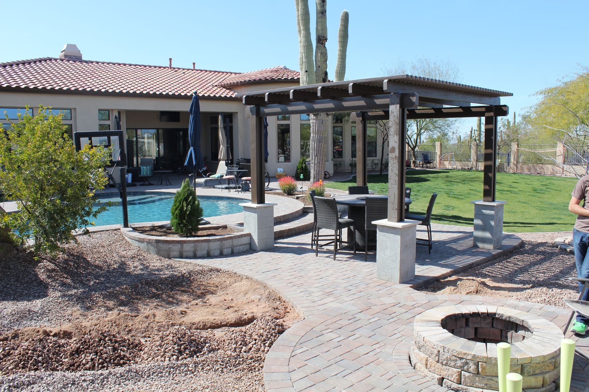 A man is standing in front of a large house with a pergola and a fire pit