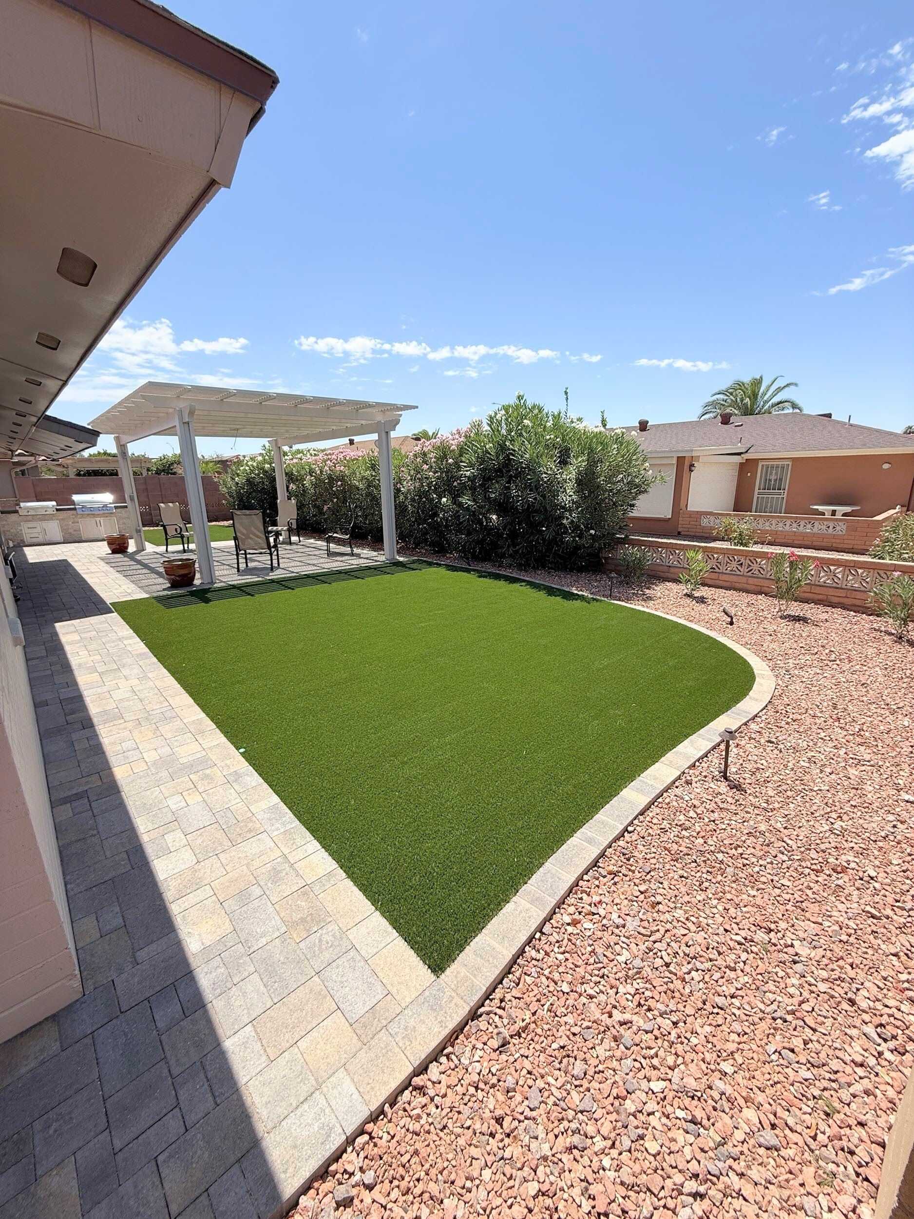 Backyard with artificial turf, pergola, patio, and red gravel landscaping on a sunny day.