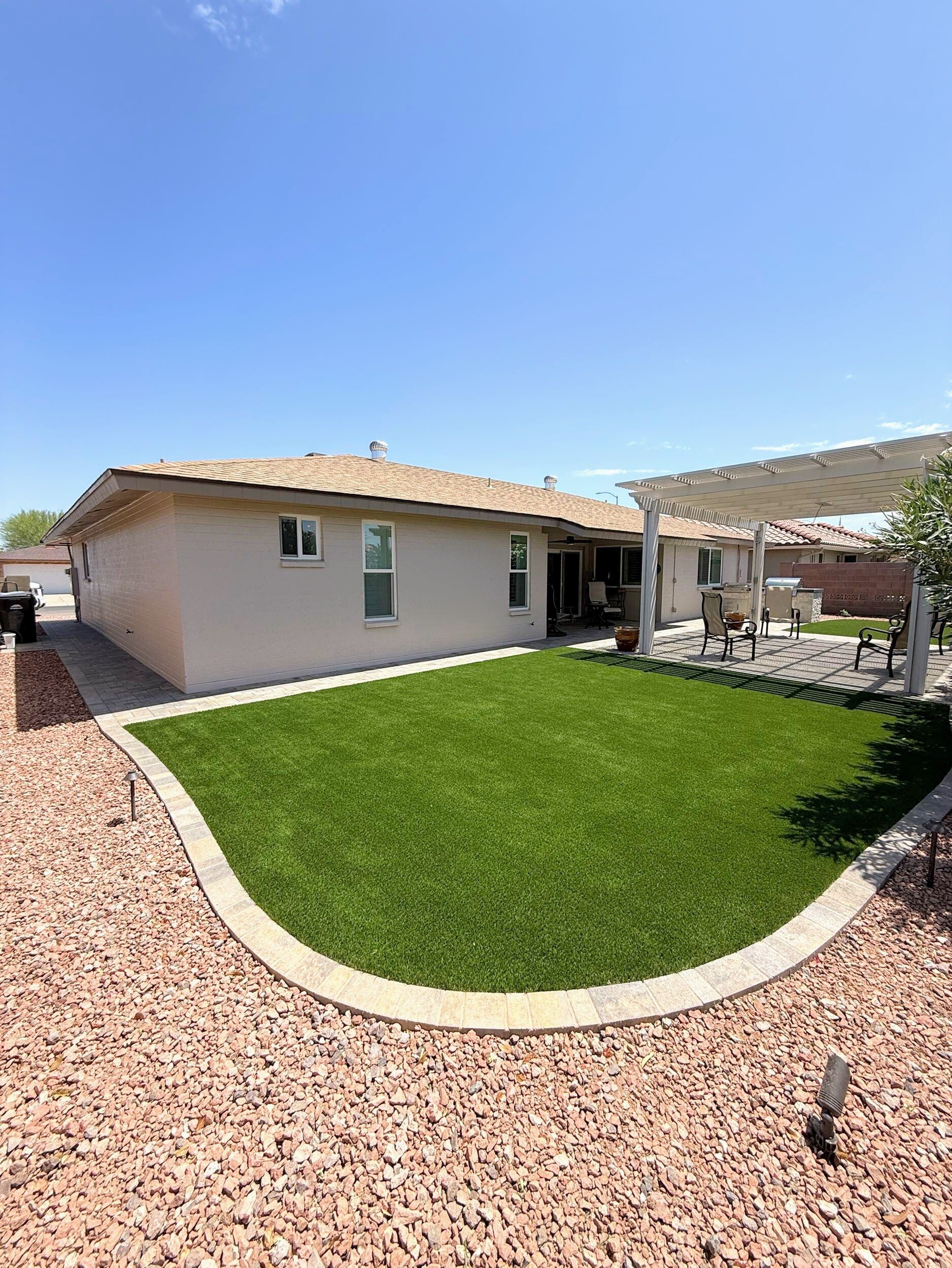 Backyard with green turf, red rock border, beige house, and pergola under a blue sky.