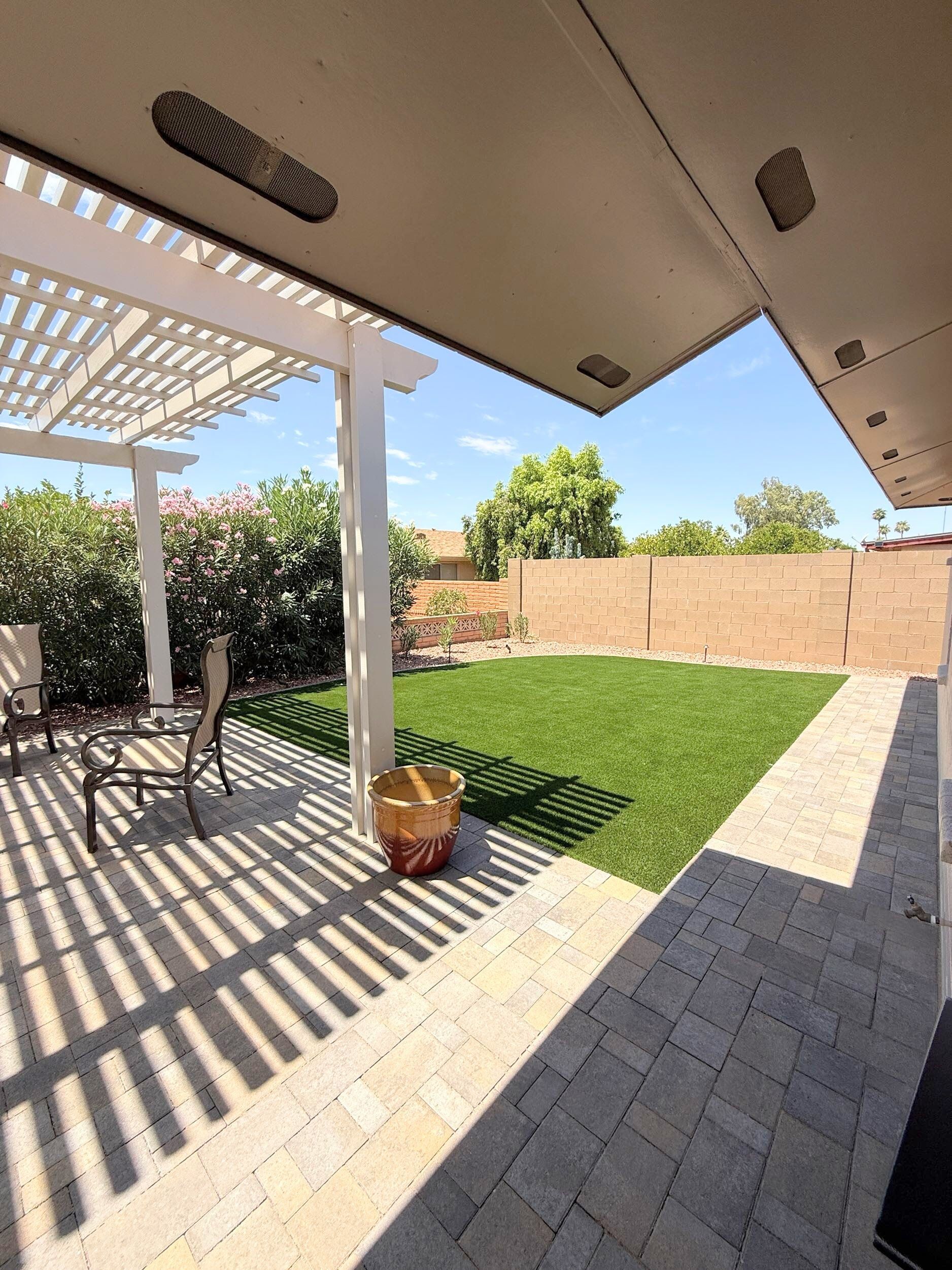 Patio with pergola and brick paving, overlooking a green lawn and tan fence on a sunny day.