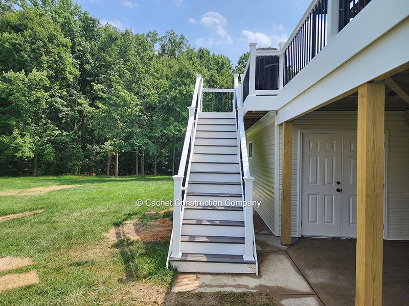Outdoor staircase leading to a deck; stairs are white and gray, set against a green yard and a building.