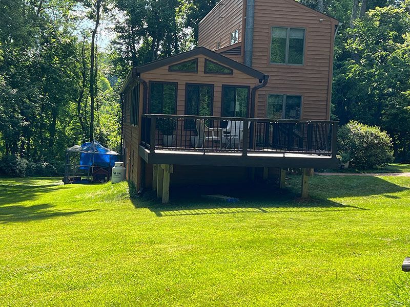 Brown house with a deck, surrounded by green grass and trees on a sunny day.