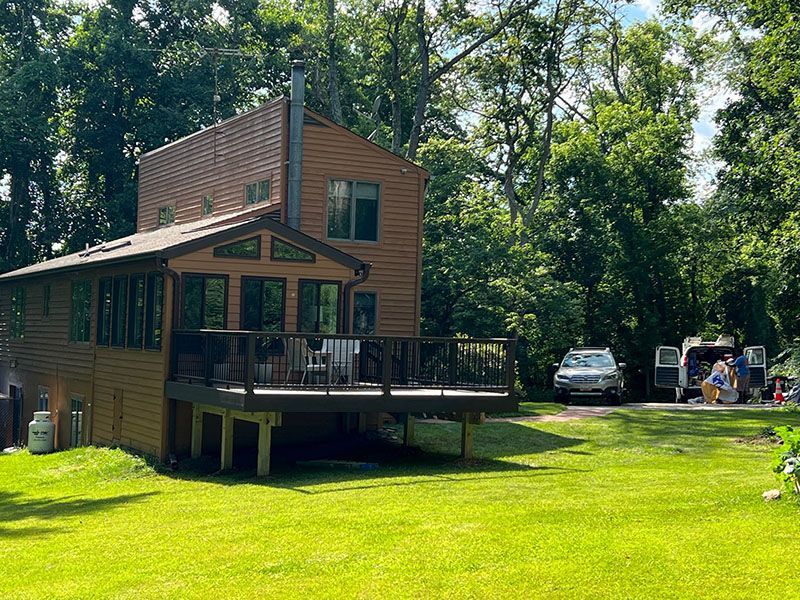 Two-story cabin with a deck, surrounded by trees, and a car with people near it.