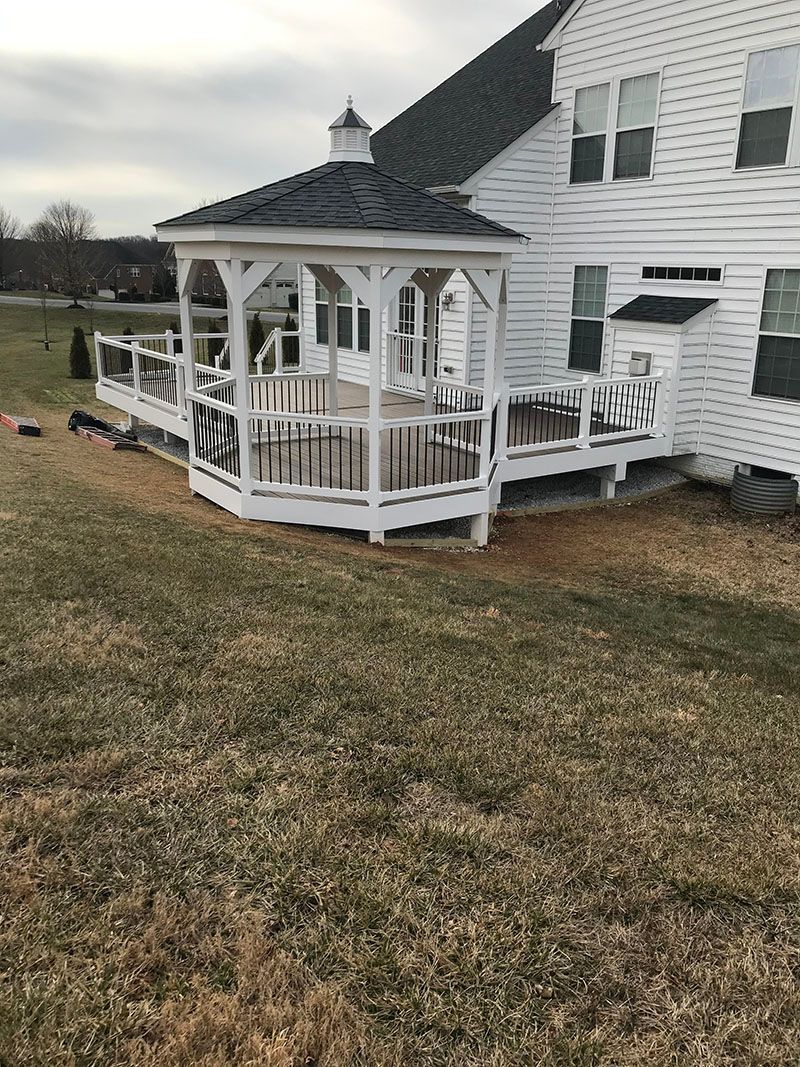 White gazebo with deck attached to a white house, set on a grassy lawn under an overcast sky.