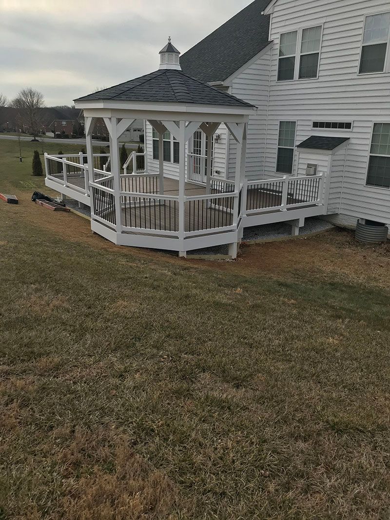 White gazebo with a dark roof on a white deck attached to a white house, surrounded by grass.