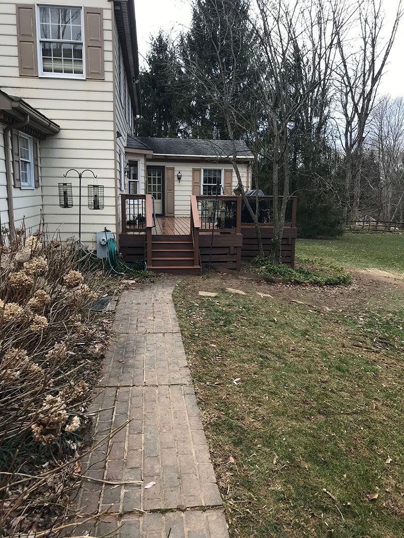 Brick pathway leads to a wooden deck and small house. Overgrown landscaping and a two-story house visible.