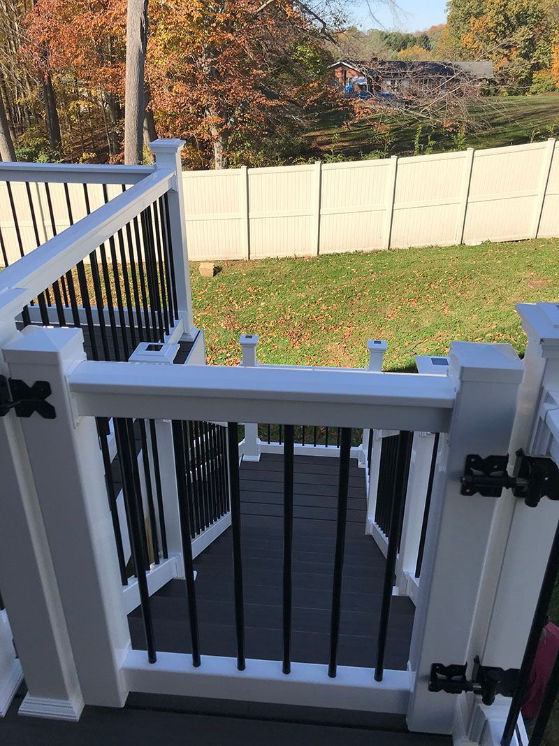 White-railed deck gate with black vertical bars, opens onto a grassy backyard with a white fence.