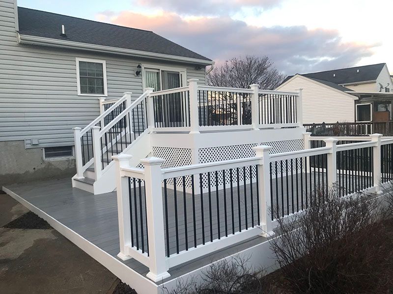 Two-level deck with white railings, black balusters, attached to a gray-sided house under a cloudy sky.