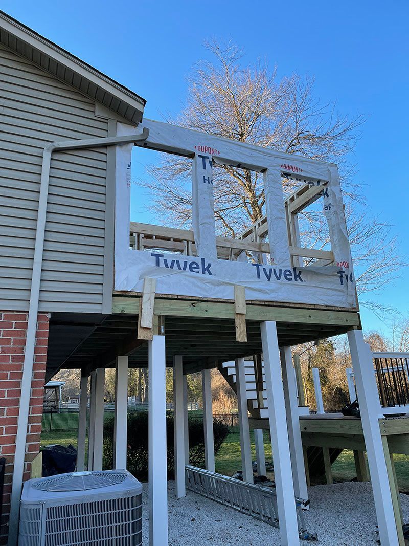Deck construction with new framing, Tyvek wrap, and supports. Side of a house with a blue sky.