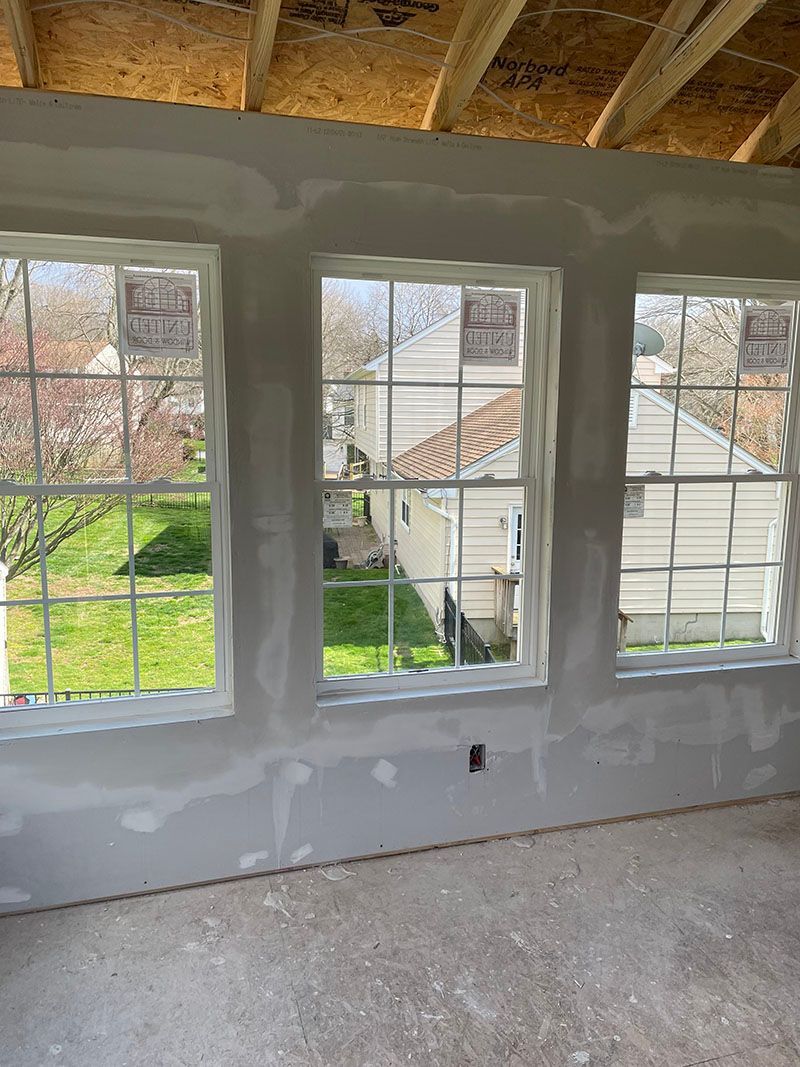 Interior view of a room with three windows, drywall walls, and exposed wooden beams.