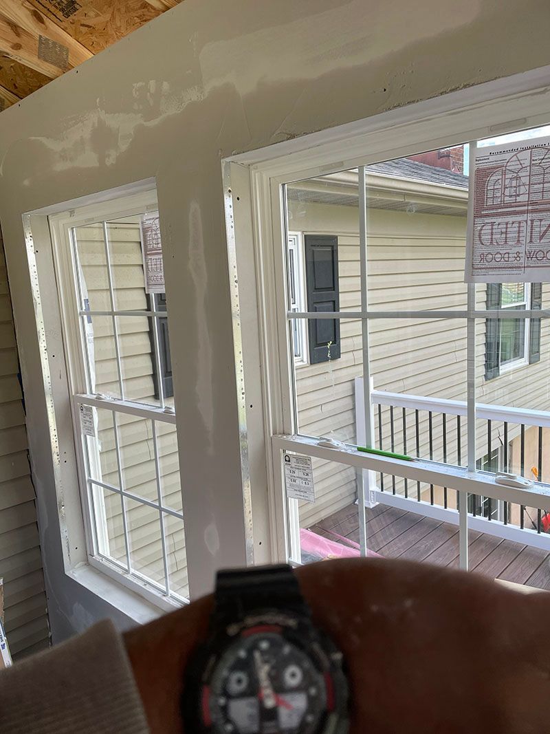 Person holding a watch in front of two white-framed windows, a house in the background. Construction site setting.
