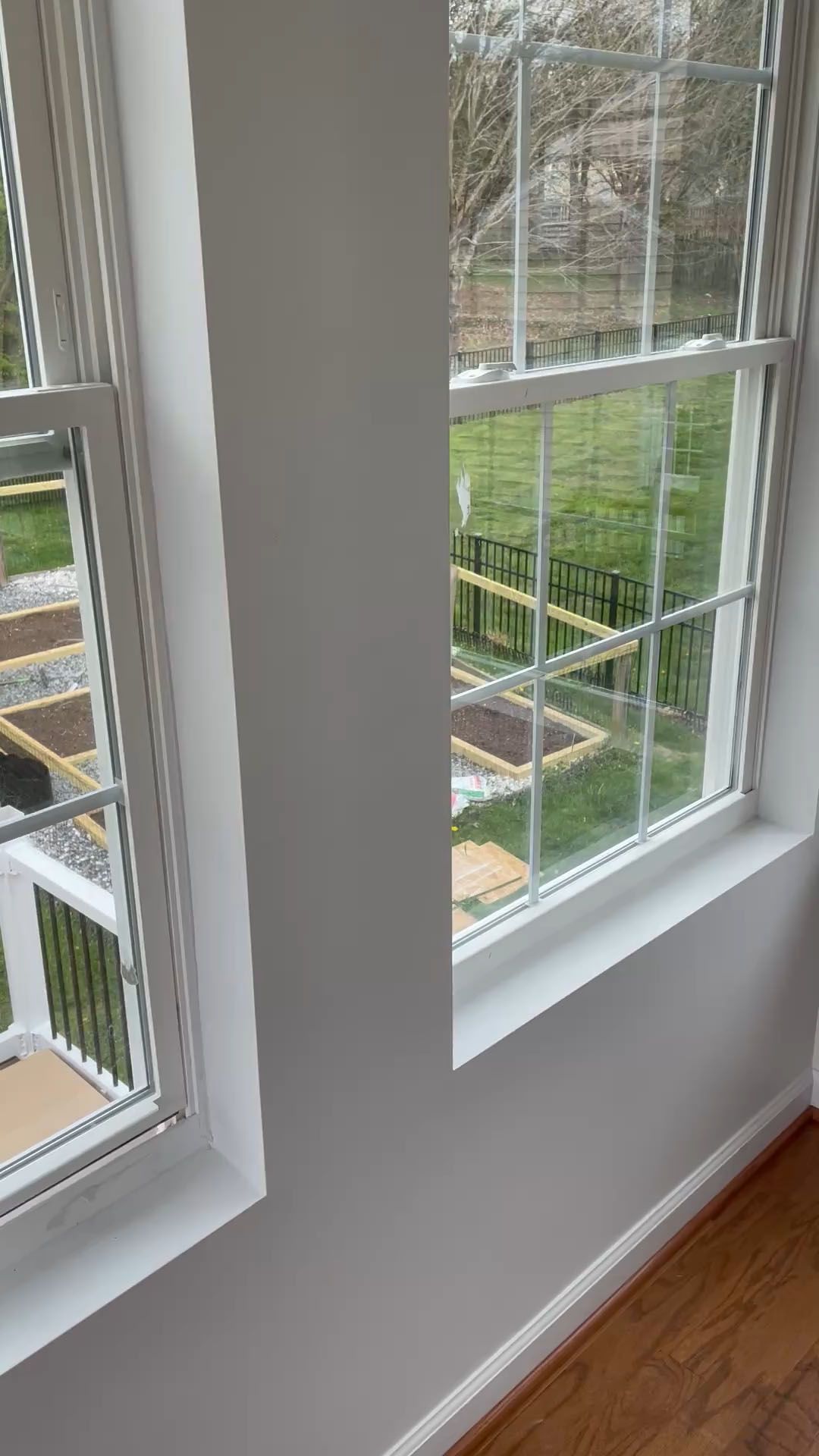 Two white-framed windows with a view of a backyard. White wall between them. Wooden floor.