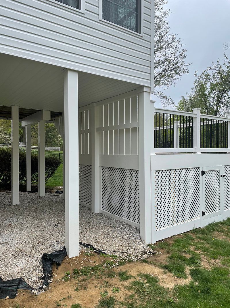 White deck with skirting. Gravel ground, white posts, and a glimpse of foliage.