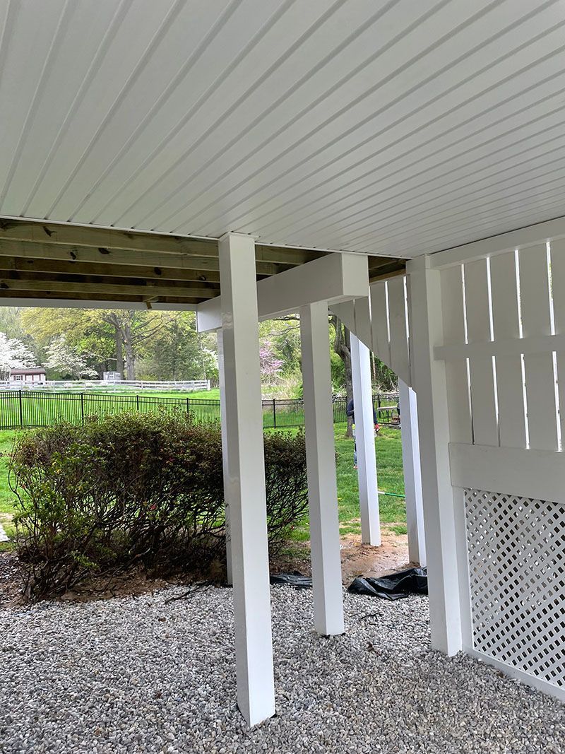 White wooden posts supporting a deck with slatted railing, set above a bed of gravel, with greenery in the background.