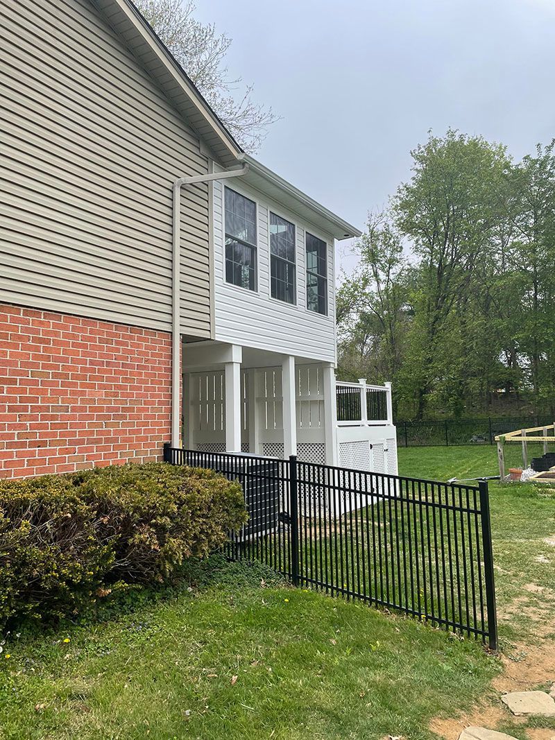 Side view of a two-story house with a porch and black fence. Grass in front, trees in the background.