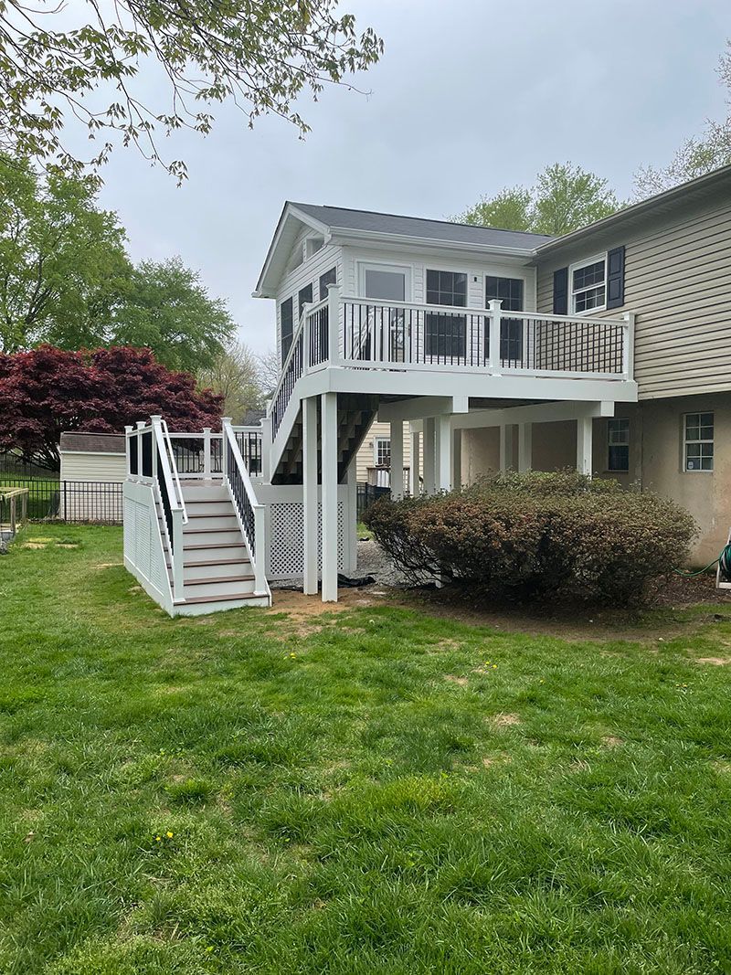 White deck with stairs attached to a house, overgrown bush, green grass, overcast sky.