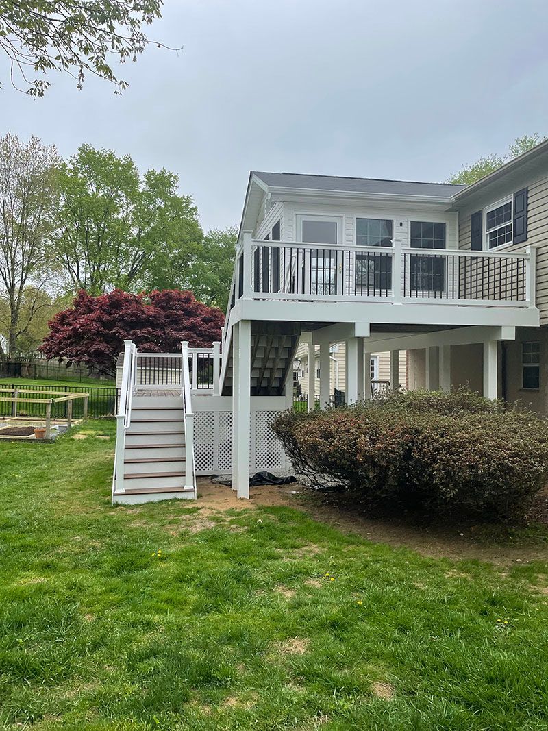Two-story deck attached to a brick house. White railing, gray stairs. Lush green lawn, cloudy sky.