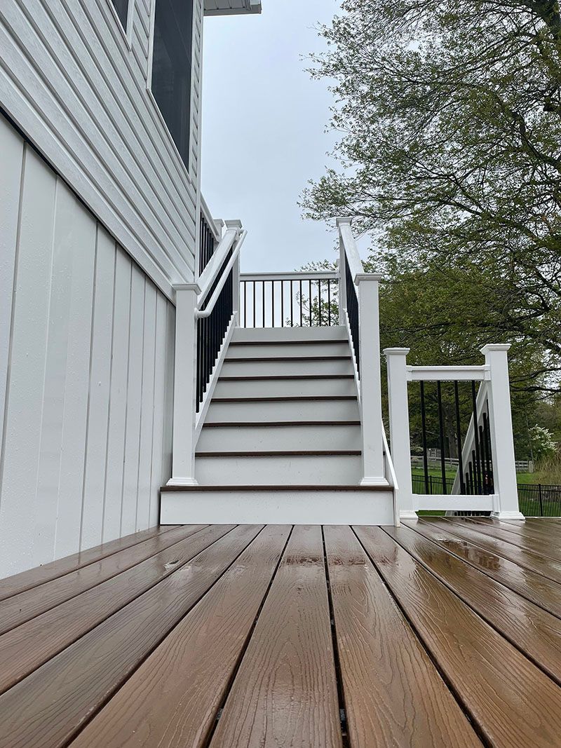 Brown deck leads to white stairs with black railing, beside white house siding.