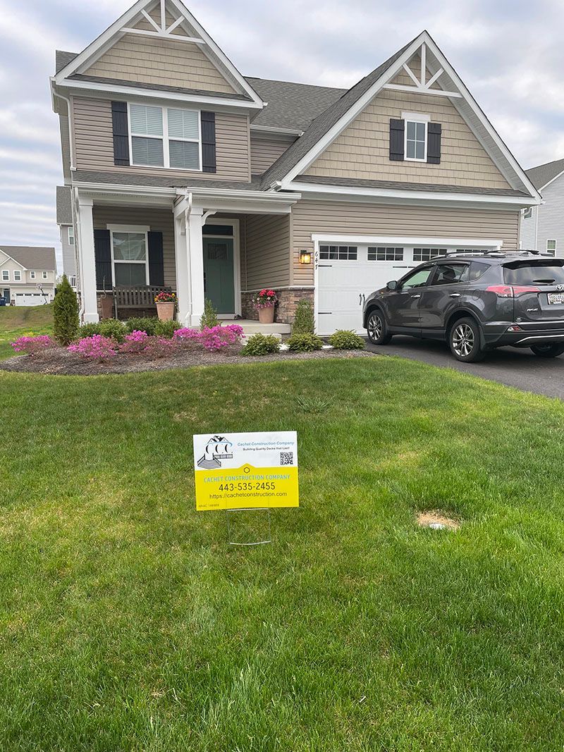 House with tan siding, black shutters, and a yellow sign on the lawn. A car is parked in the driveway.