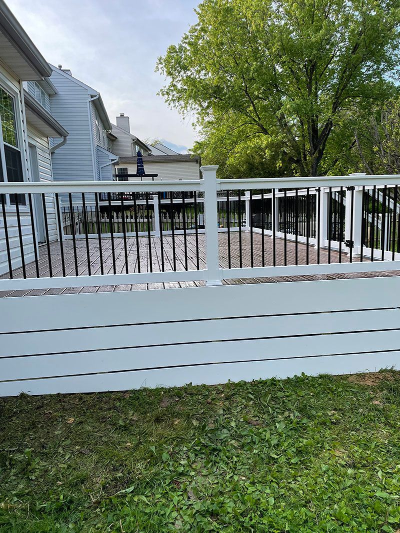 White and black deck with black railing, in front of a light blue house with a tree in the background.